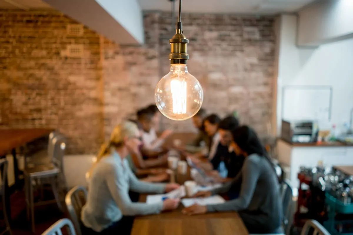 A glowing pendant bulb hanging above a group working along a long wooden table in a brick walled room