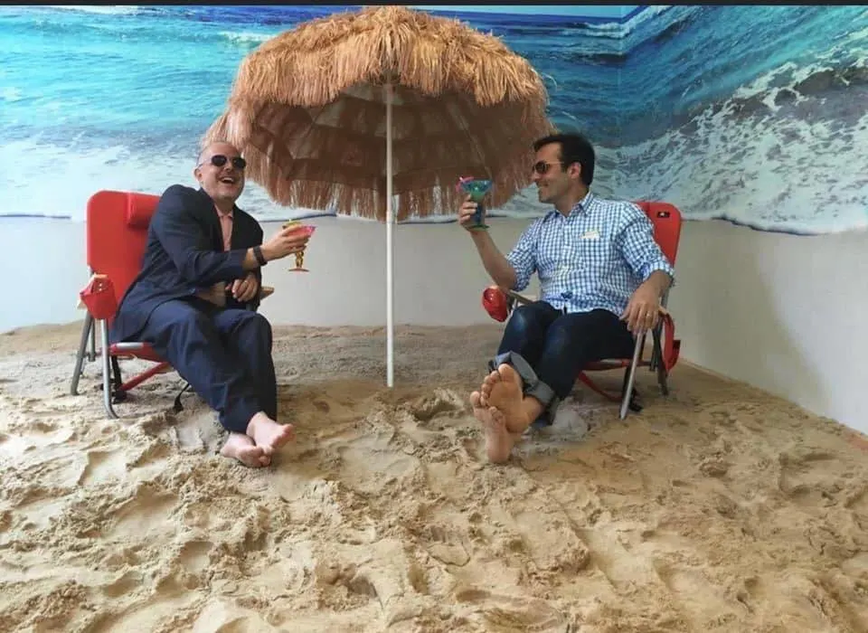 Two people in office clothes sitting barefoot with cocktails under a straw parasol on an indoor sand beach setup