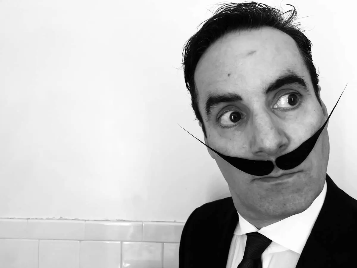 Black and white close-up of a person in a suit and tie wearing an oversized curled Dali style prop moustache