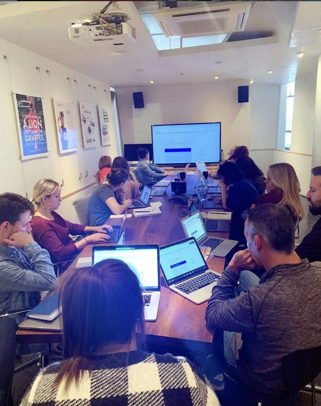 About a dozen people with laptops around a long conference table facing a wall screen in a meeting room