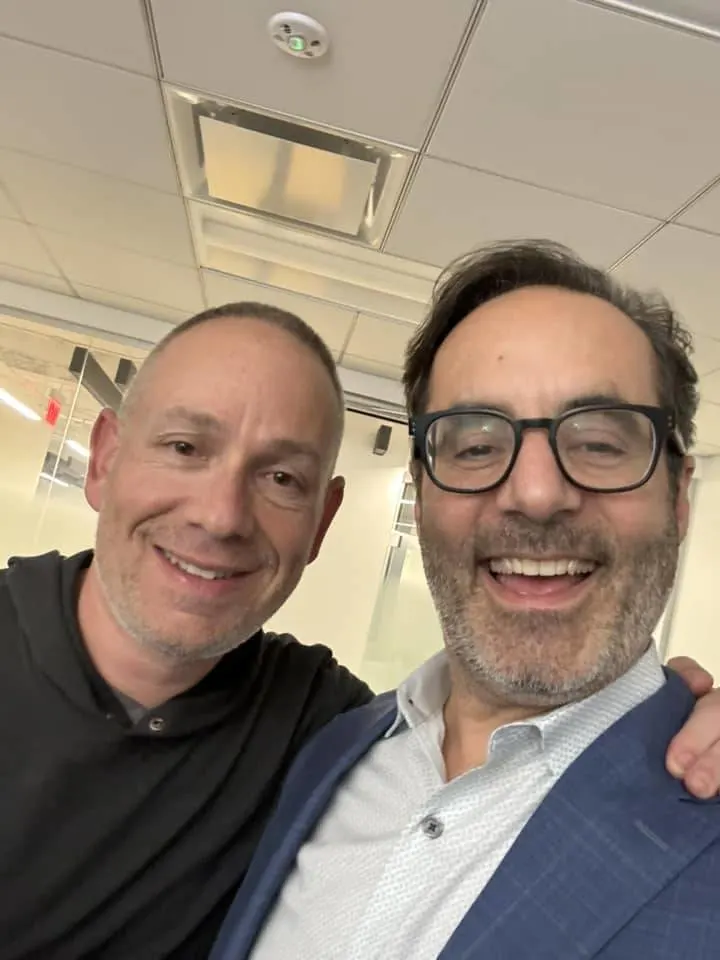 Selfie of two people smiling in an office, one in a black polo and one in glasses and a blue blazer
