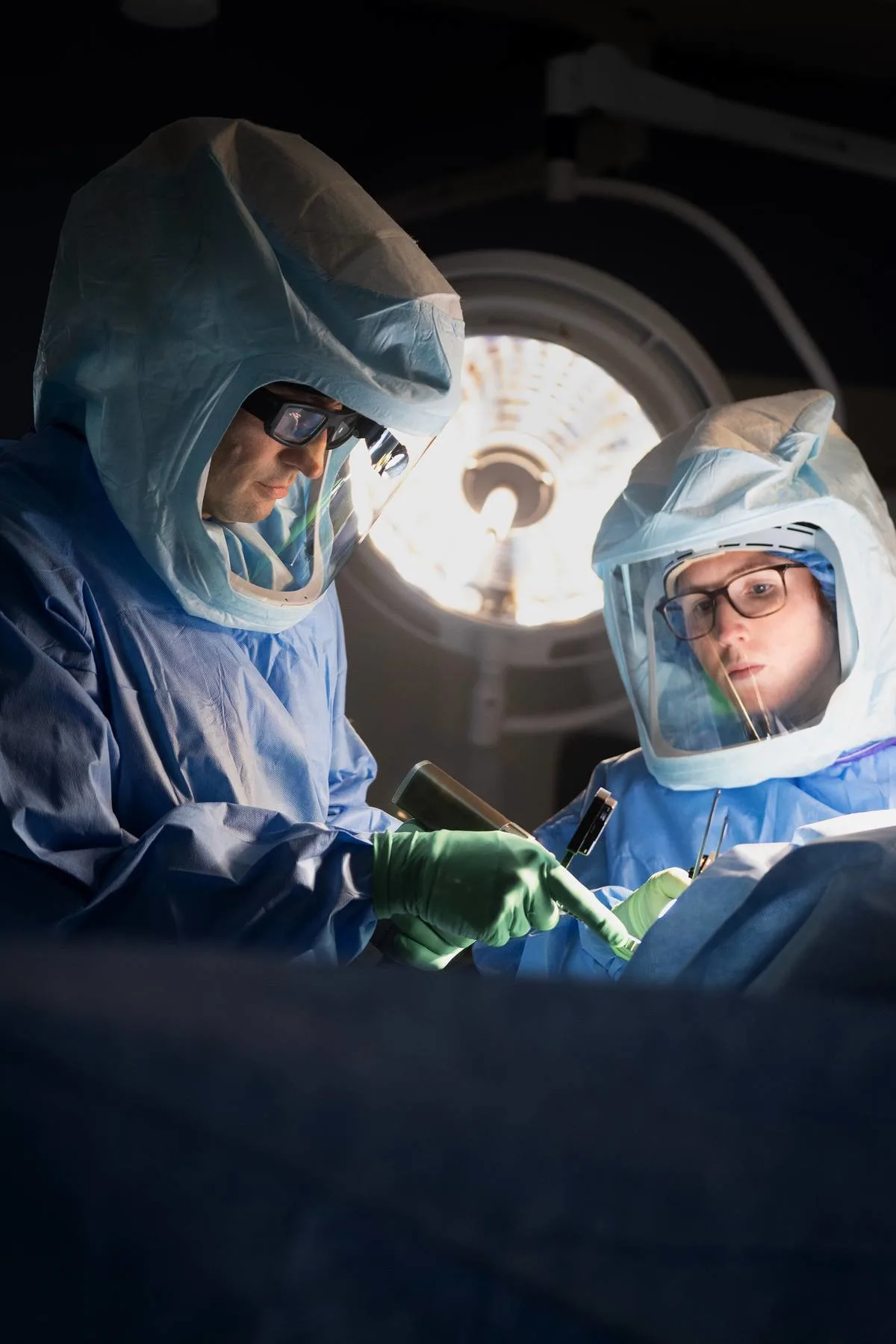 Two people in surgical hoods and green gloves working over a draped patient under an operating light
