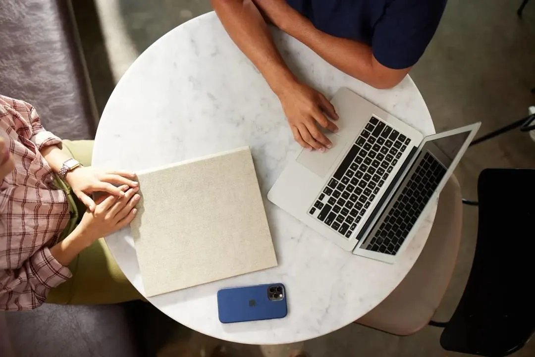 Overhead view of two people at a small round marble table, one typing on a laptop, the other with a notebook and phone